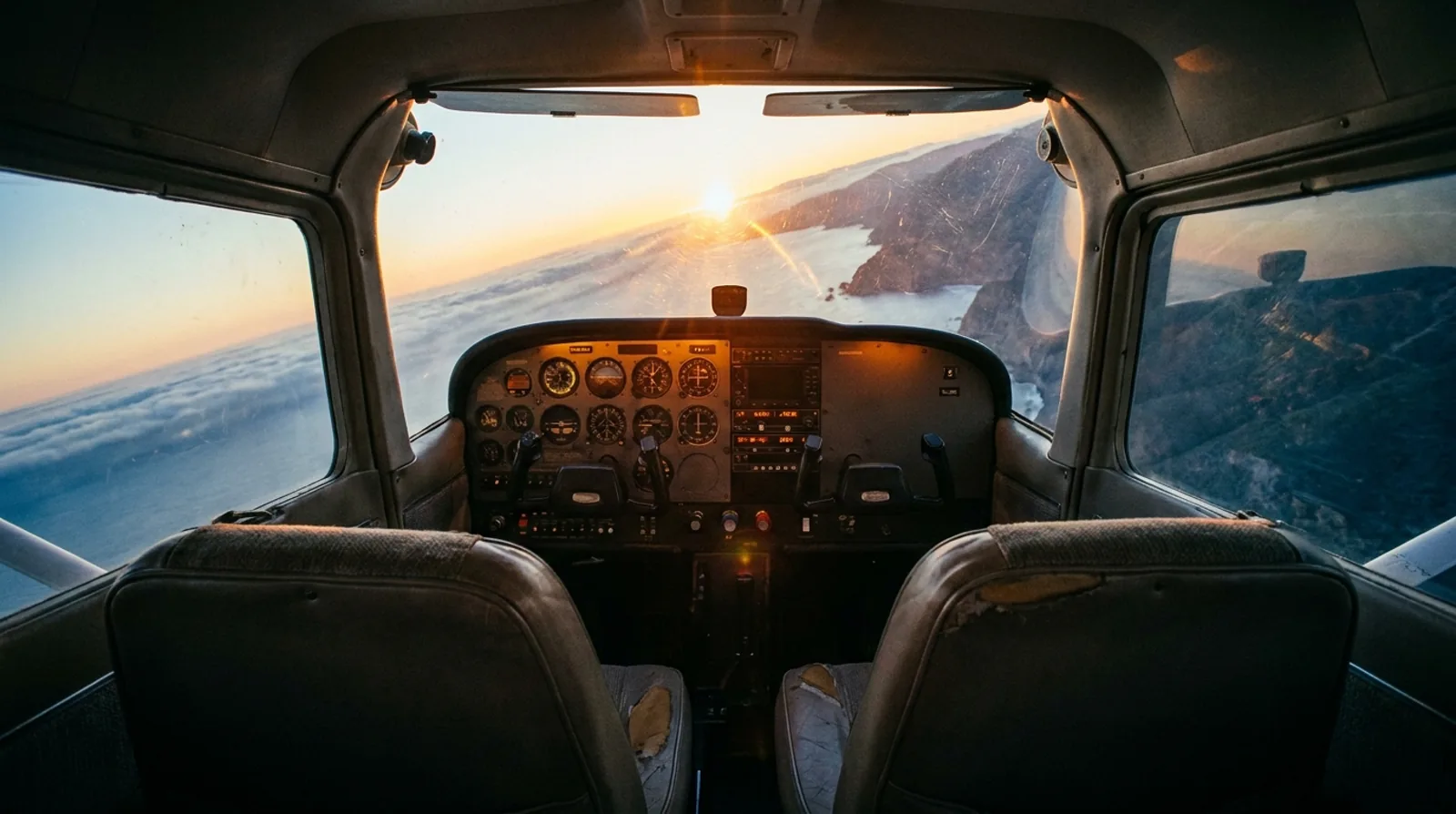 Vista desde los asientos traseros de una avioneta en vuelo al atardecer: panel de instrumentos analógicos iluminado y costa bajo una capa de nubes